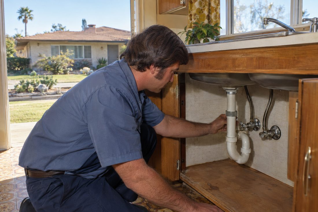 Close view of a plumber inspecting the shutoff connections beneath a kitchen sink in a 1970s Vista Village ranch home in Vista Village, CA